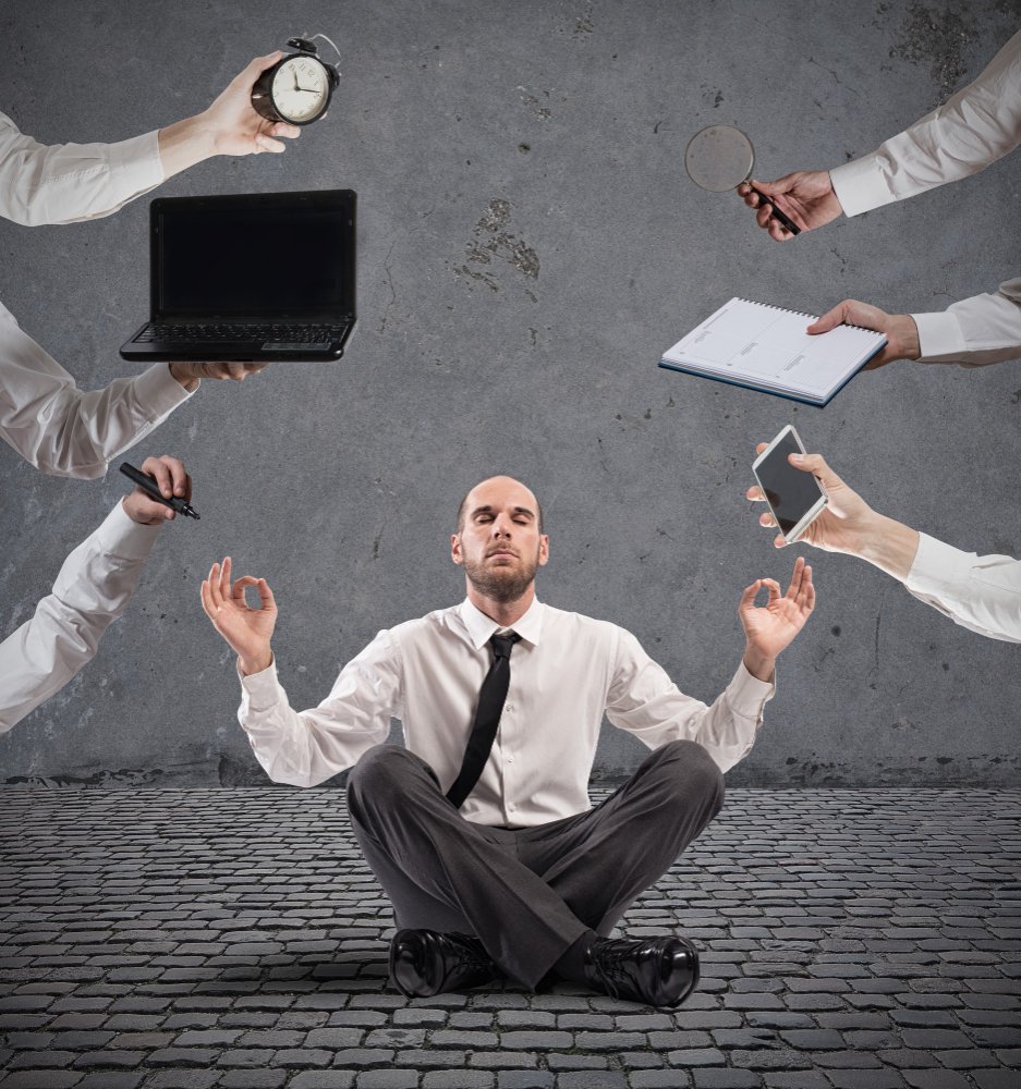 Businessman meditating calmly on a cobblestone floor while multiple hands offer distractions like a phone, laptop, clock, and notepad—symbolizing the need for longevity strategies in a high-pressure lifestyle.