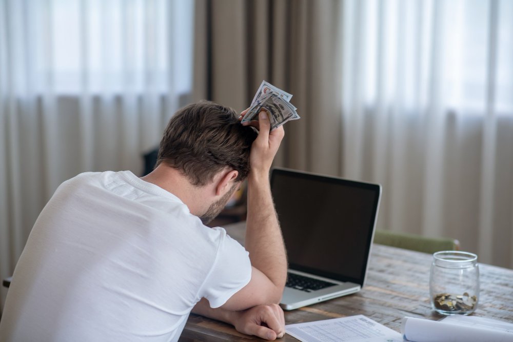 Frustrated man holding dollar bills while sitting in front of a laptop, appearing stressed about finances, with documents and a coin jar on the table.